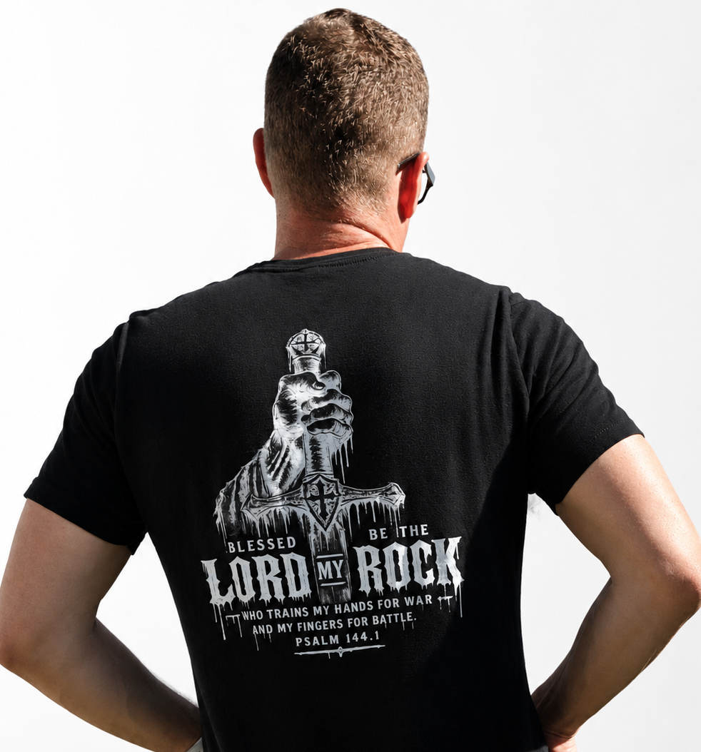 A man viewed from behind wearing a black t-shirt with a large white graphic print on the back. The graphic features a soldier statue and the text 'BLESSED BE THE LORD MY ROCK' with a Bible verse below.