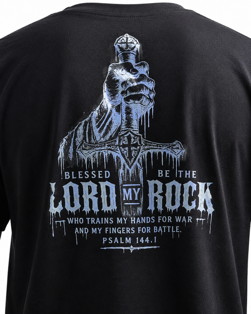 Close-up of the back of a black short-sleeve T-shirt featuring a large distressed graphic print in light blue and white. The design shows a crowned skull-like figure above a sword and text reading 'Blessed Be the Lord My Rock Who Trains My Hands for War and My Fingers for Battle.