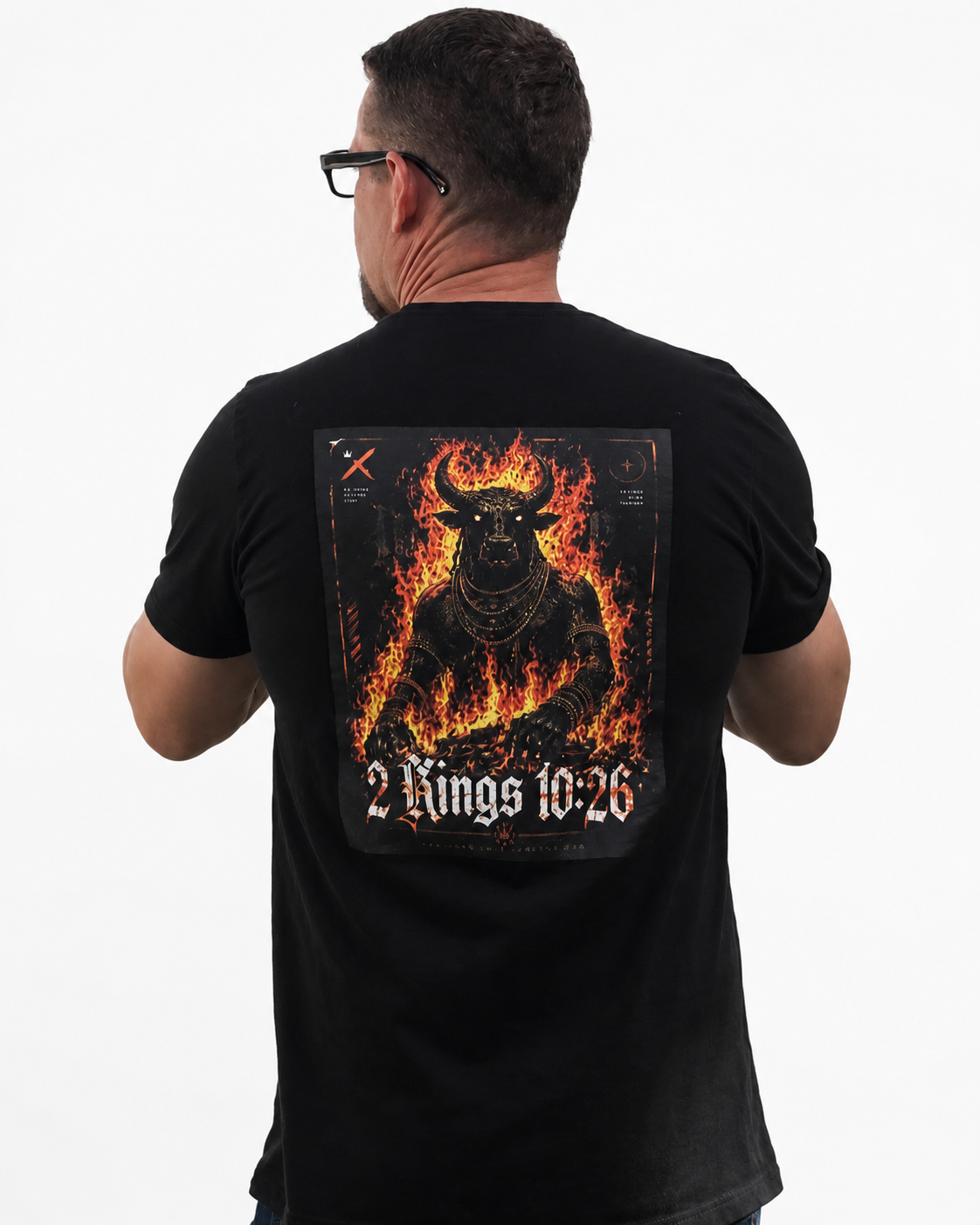 A man viewed from behind wearing a black t-shirt with a large graphic print on the back. The graphic features a fiery, horned demon figure in orange and yellow flames, with the text '2 Kings 10:26' in a gothic font at the bottom.