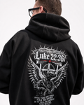 A man wearing a black hoodie, viewed from the back. The hoodie features a large white graphic print on the back with the text 'Luke 22:36' and an illustration of a winged soldier holding a sword.