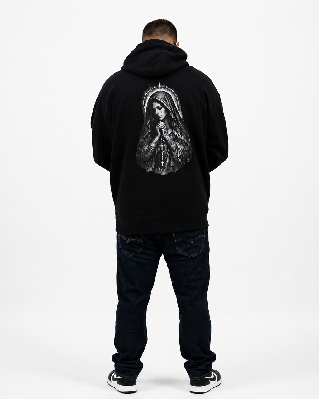 A man viewed from behind wearing a black pullover hoodie with a large white graphic of the Virgin Mary on the back, paired with dark jeans and black-and-white sneakers, against a plain white studio background.