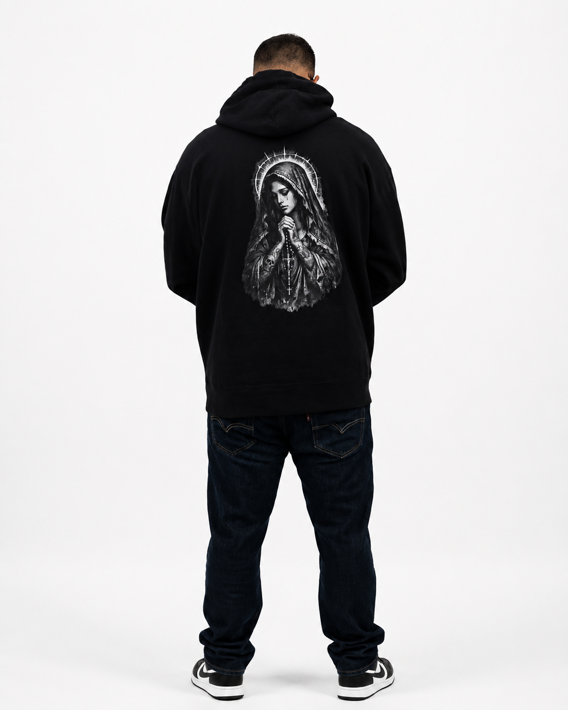 A man viewed from behind wearing a black pullover hoodie with a large white graphic of the Virgin Mary on the back, paired with dark jeans and black-and-white sneakers, against a plain white studio background.